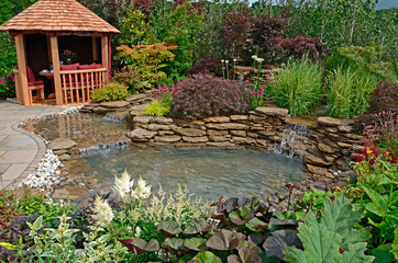 Fototapeta na wymiar The pond area and terrace with Summer House in an aquatic garden with planted rockery and outdoor eating and selection of flowers, shrubs and grasses