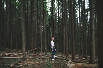 Naklejka na ściany i meble Young caucasian tourist girl in white T-shirt walking in the mountain fir forest feeling relaxed and calm. Attractive female hiker looking at camera standing in the middle of the coniferous woods.