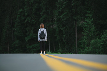Horizontal shot of female traveler walking down the road at evening mountain forest background. Tourist girl with a backpack in the middle of the asphalt road near the woods.