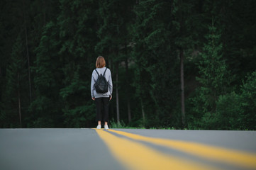 Obraz premium Horizontal shot of female traveler walking down the road at evening mountain forest background. Tourist girl with a backpack in the middle of the asphalt road near the woods.