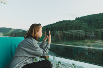 Young female traveler in grey sweatshirt taking photo on smartphone of mountain forest landscape sitting on the restaurant terrace. Shot of tourist woman using phone to photograph fir woods and lake.