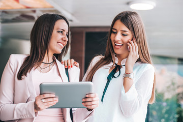 Two attractive young women looking at the tablet screen together and talking on mobile phone