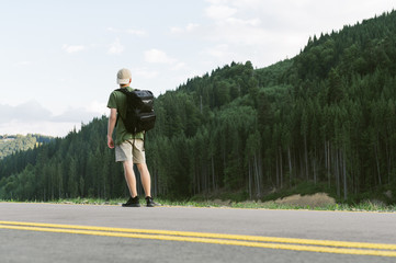Fototapeta premium Full length photo of a young tourist guy standing on the road looking at beautiful mountain forests scenery. Back view shot of male traveler with backpack enjoying coniferous forests and mountains.