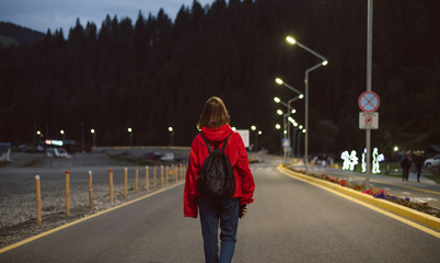 Female traveler in a red raincoat walking down the road in the mountainous scenery in the evening. Young tourist girl back view on the road going to the mountain forest in the street lights.