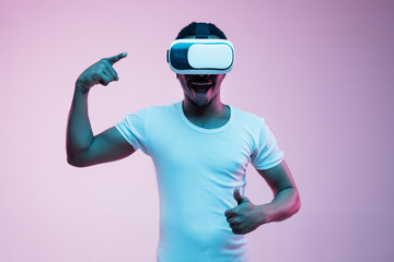 Young african-american man's playing in VR-glasses in neon light on gradient background. Male portrait. Concept of human emotions, facial expression, modern gadgets and technologies. Pointing in game.