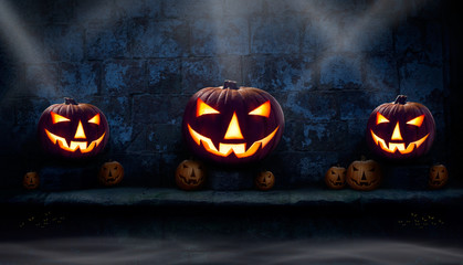 Three lit halloween Jack O Lanterns on the left, right and centre on a stone plinth  at night with many unlit lanterns and smoke surrounding them.