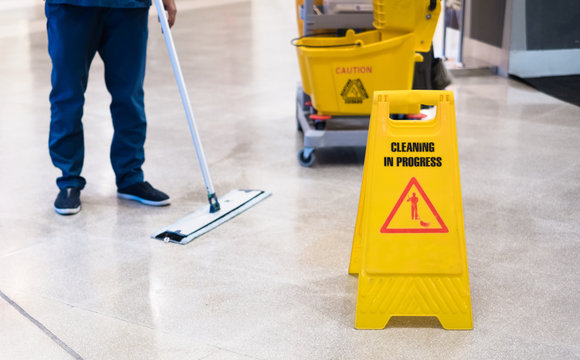 Janitor Cleaning Floor In Front Of Yellow Caution "Cleaning in progress". Cleaning service in public area.