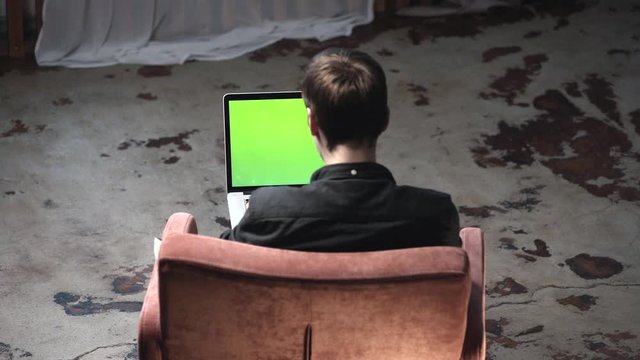 View from the back of young man in black shirt sitting in the brown comfortable chair and working on laptop computer with chroma key green screen. Stock footage. Place of your own content.