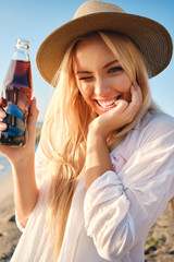 Young joyful blond woman in hat holding bottle of soda water happily winking while looking in...