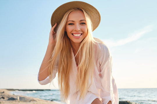 Young attractive smiling blond woman in shirt and hat joyfully looking in camera with sea on background