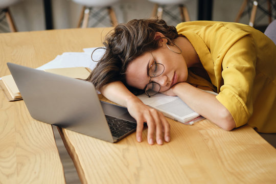 Young tired woman in eyeglasses asleep on desk with laptop and documents under head at workplace