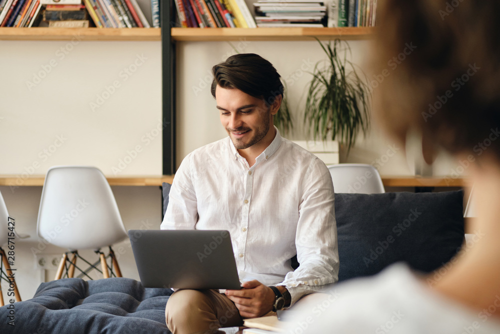 Poster young handsome smiling businessman sitting on sofa with colleague while happily working on laptop in