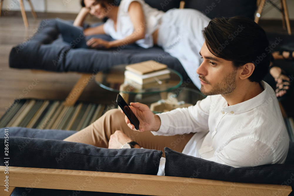 Poster side view of young businessman sitting on sofa with cellphone thoughtfully looking aside in co-worki