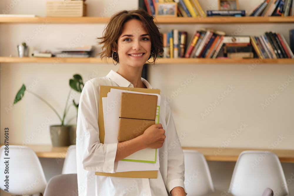 Poster young attractive smiling woman joyfully looking in camera with papers and notepad in office