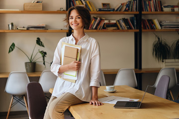 Young cheerful woman in white shirt joyfully looking in camera while sitting on desk with papers...