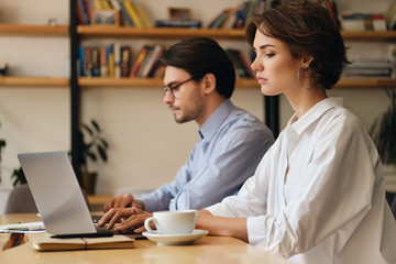 Young serious woman sitting at the table tiredly working on laptop with colleague near in office