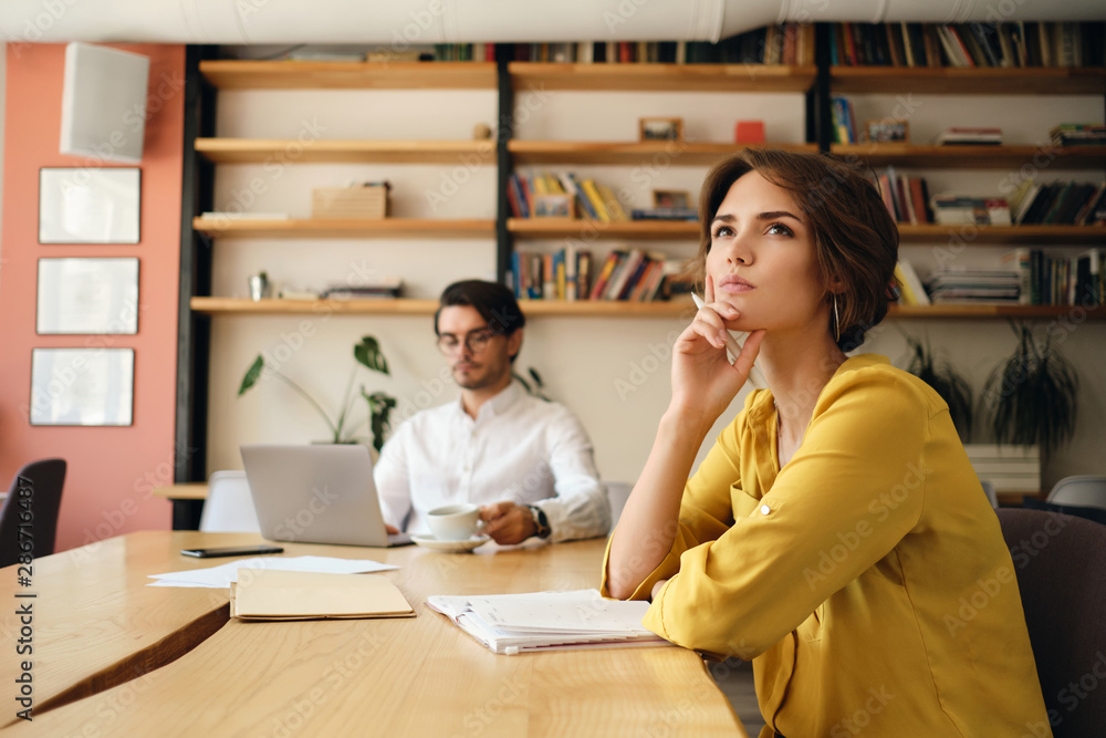 Aufkleber Young pensive woman sitting at the table with notepad dreamily looking aside while working with colleague on background in modern office