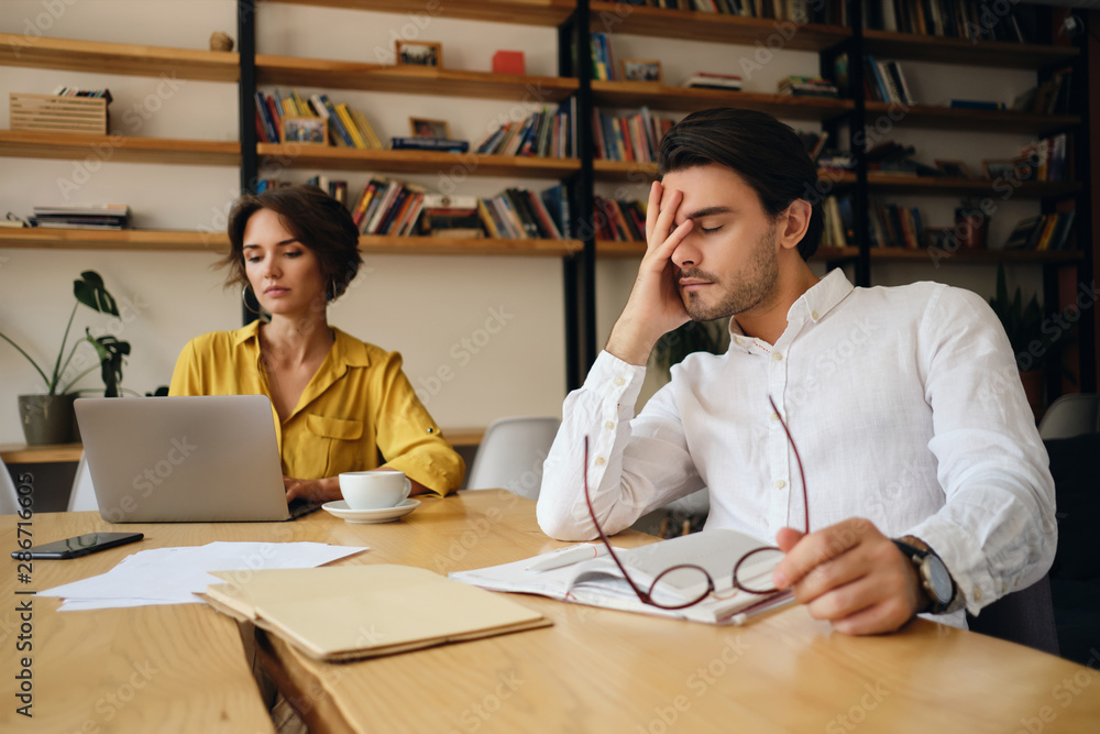 Poster young exhausted man sitting at the table with notepad and eyeglasses while tiredly holding hand near