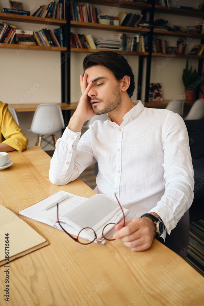 Poster young tired man sitting at the table with notepad leaning head on hand sleeping at work in modern of
