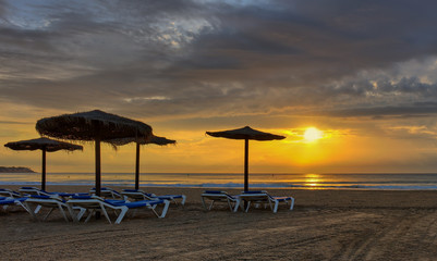 A dream beach with umbrellas made of straw and blue beach chairs in the early morning at sunrise on the Mediterranean sea. Beautiful clouds adorn the sky.