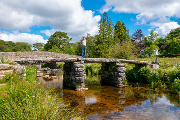 Fototapeta na wymiar Senior male tourist standing on ancient clapper bridge at Postbridge, Dartmoor National Park, Devon, England