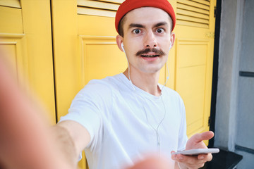 Young attractive mustache man in T-shirt and red hat with cellphone while amazedly taking photo on...