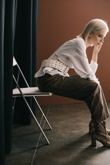 side view of stylish blonde woman in white blouse and boots with snakeskin print sitting on chair...