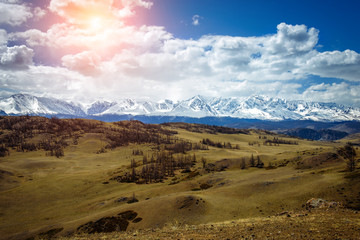 Amazing mountain landscape. Rocky mountains with snowy peaks, hills covered with grass in the Alpine scene on a bright sunny day with blue sky and clouds. View of steppe and snow-covered mountains.