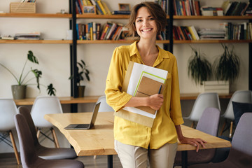 Young cheerful woman in yellow shirt leaning on desk with notepad and papers in hand while joyfully...