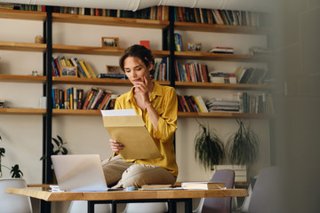 Young attractive woman in yellow shirt sitting on desk with laptop while dreamily working with...