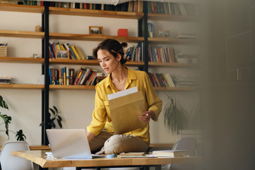 Young beautiful pensive woman in yellow shirt sitting on desk with papers while dreamily working on...