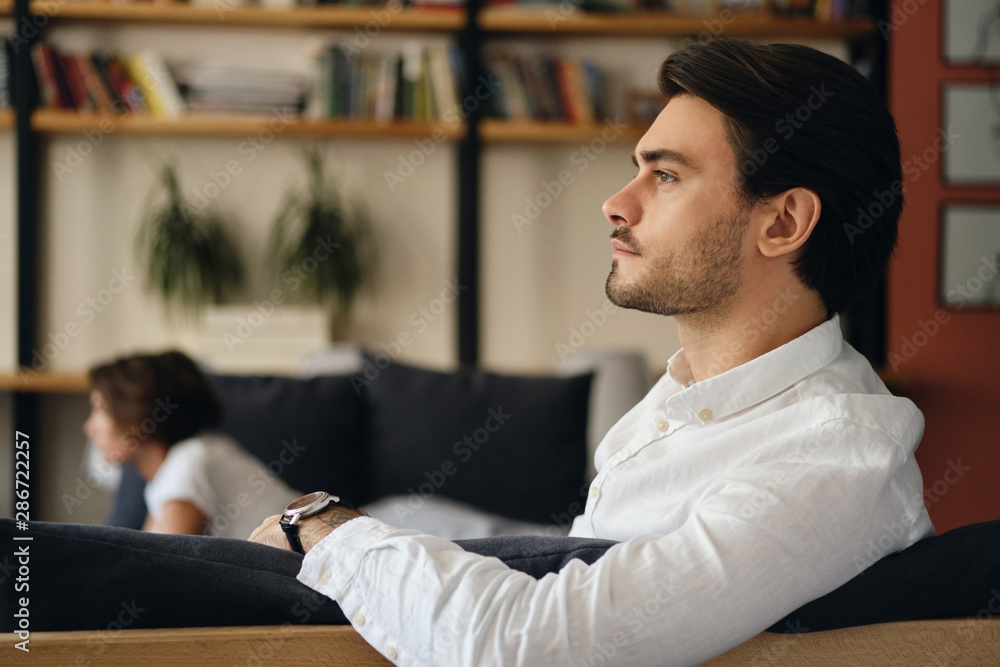 Poster side view of young handsome businessman sitting on sofa with colleague while thoughtfully looking as