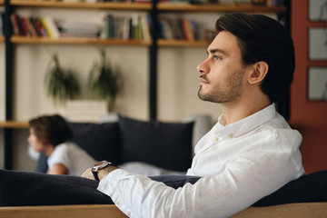 Side view of young handsome businessman sitting on sofa with colleague while thoughtfully looking aside in modern co-working space