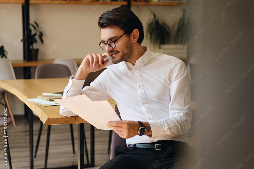 Poster young attractive smiling businessman in eyeglasses happily reading papers while working in modern of