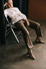 stylish blonde woman in white blouse and boots with snakeskin print sitting on chair near curtain...