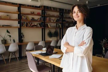 Fotobehang Young attractive woman in white shirt dreamily looking in camera with desk on background in office © Anton