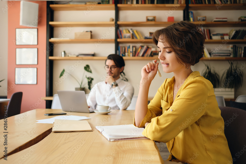 Poster young attractive woman sitting at the table with notepad and dreamily working with colleague on back