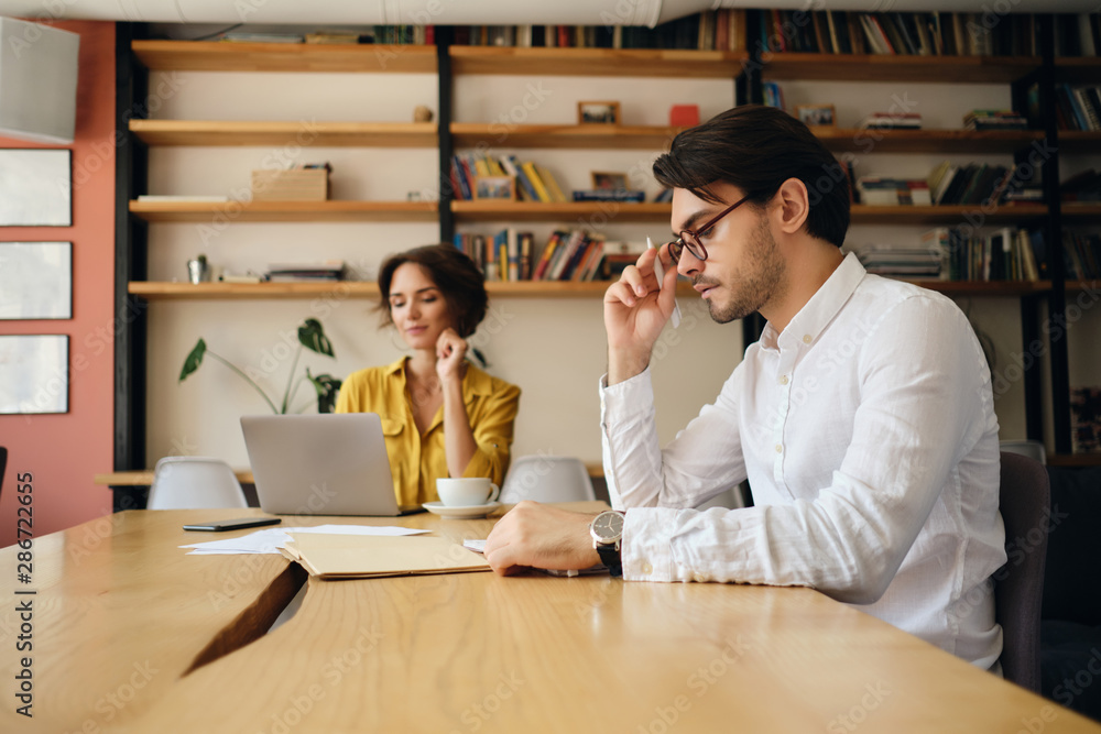 Poster young handsome man in eyeglasses sitting at the table thoughtfully working with colleague on backgro