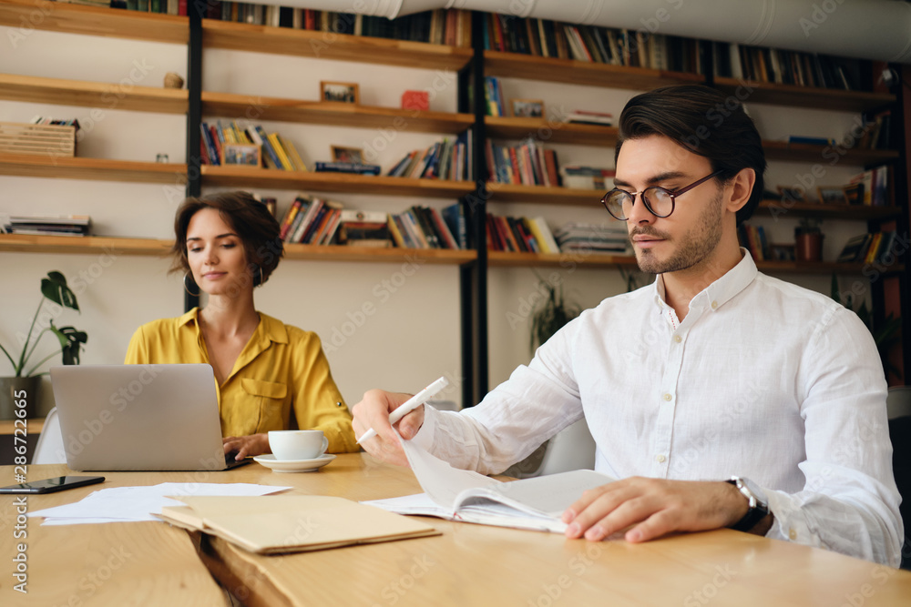 Poster young serious man in eyeglasses sitting at the table thoughtfully working in modern office with coll