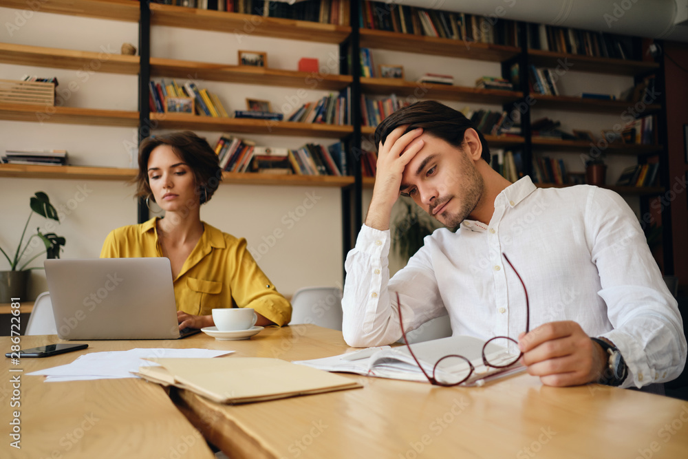 Aufkleber Young tired man sitting at the table with notepad and eyeglasses while thoughtfully holding hand near head at work in office with colleague on background