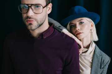 stylish young man in glasses and blonde woman in hat looking at camera near curtain
