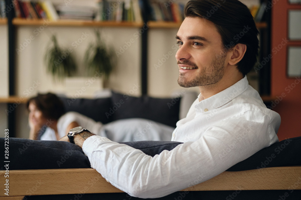 Poster young attractive smiling businessman sitting on sofa with colleague while happily looking aside in m