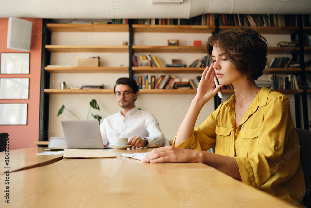 Poster young serious woman sitting at the table with notepad thoughtfully working with colleague near in of