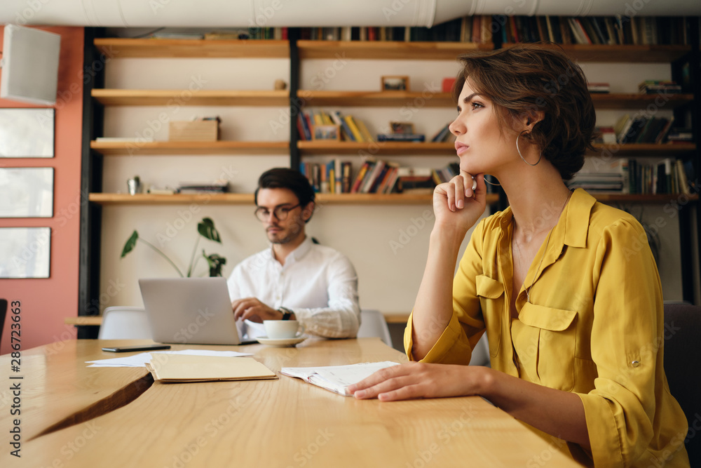 Poster side view of young woman sitting at the table with notepad thoughtfully working with colleague on ba