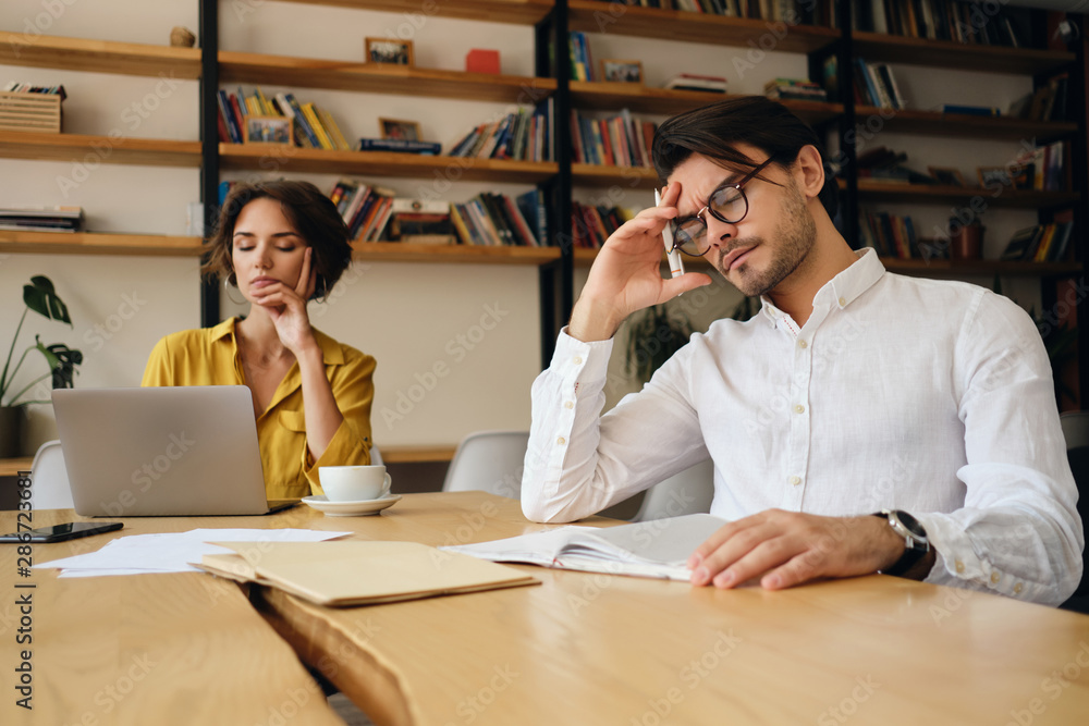 Poster young tired man in eyeglasses sitting at the table leaning head on hand while thoughtfully working i
