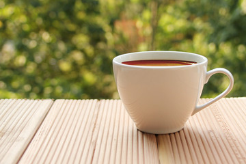 white cup with coffee on a light wooden table against a green summer garden, close-up, copy space