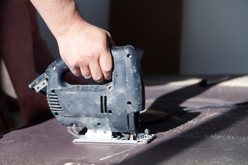 Closeup hand of woodworker with professional cutting tool fretsaw or jigsaw, cut wooden tabletop, sawing plank, brown filings, sawdust. Concept cut hole for the sink in the kitchen