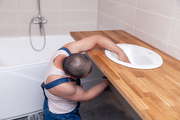 Closeup male plumber worker in blue denim uniform, overalls, fixing sink in bathroom with tile...