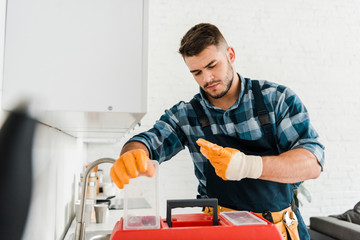 selective focus of handsome handyman looking at toolbox in kitchen