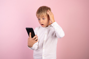 Fototapeta premium Portrait of male schoolboy isolated on pink reading articles, doing work searching for information in internet using his smartphone. Troublesome boy making research with emotional face expression